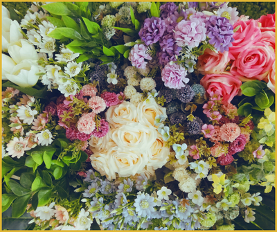 A vibrant floral arrangement featuring a mix of white roses, pink roses, lavender-hued hydrangeas, daisies, and small filler flowers in shades of cream, pink, yellow, and purple, all surrounded by lush green foliage.