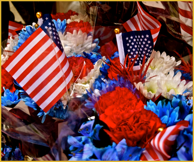 A vibrant display of patriotic floral arrangements featuring red, white, and blue flowers, such as carnations and chrysanthemums, adorned with small American flags and wrapped in clear cellophane for presentation.