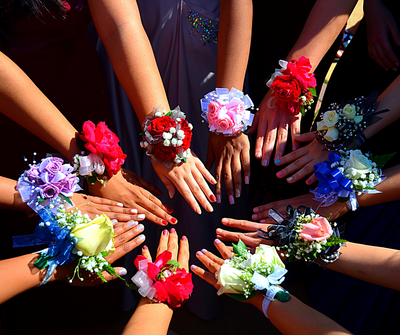 A group of individuals extending their hands in a circle, each wearing colorful wrist corsages made of roses, baby’s breath, and ribbons in various shades, including red, pink, blue, purple, white, and yellow.