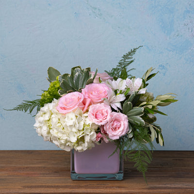 Square glass vase with pink roses, white hydrangeas, alstroemeria, and greenery in a compact floral arrangement.