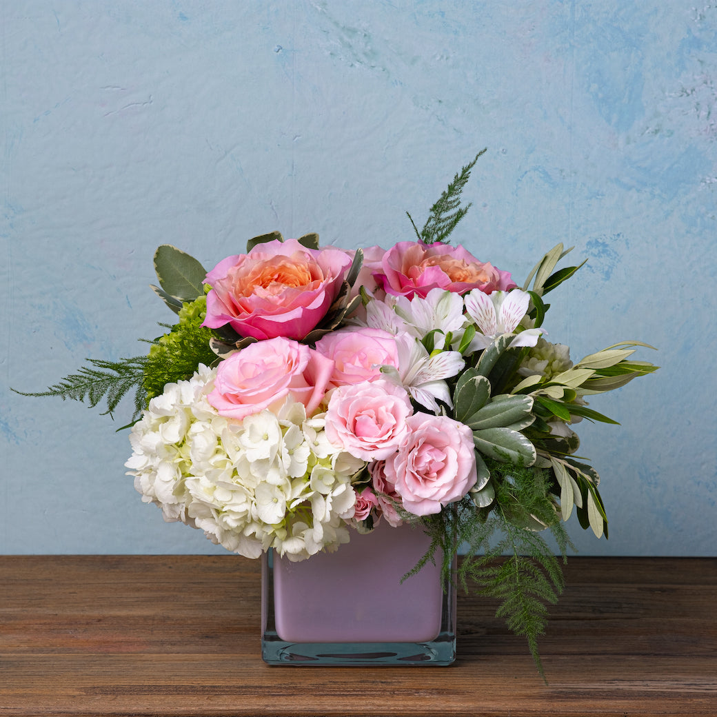 Square glass vase with pink roses, white hydrangeas, alstroemeria, and greenery in a compact floral arrangement.