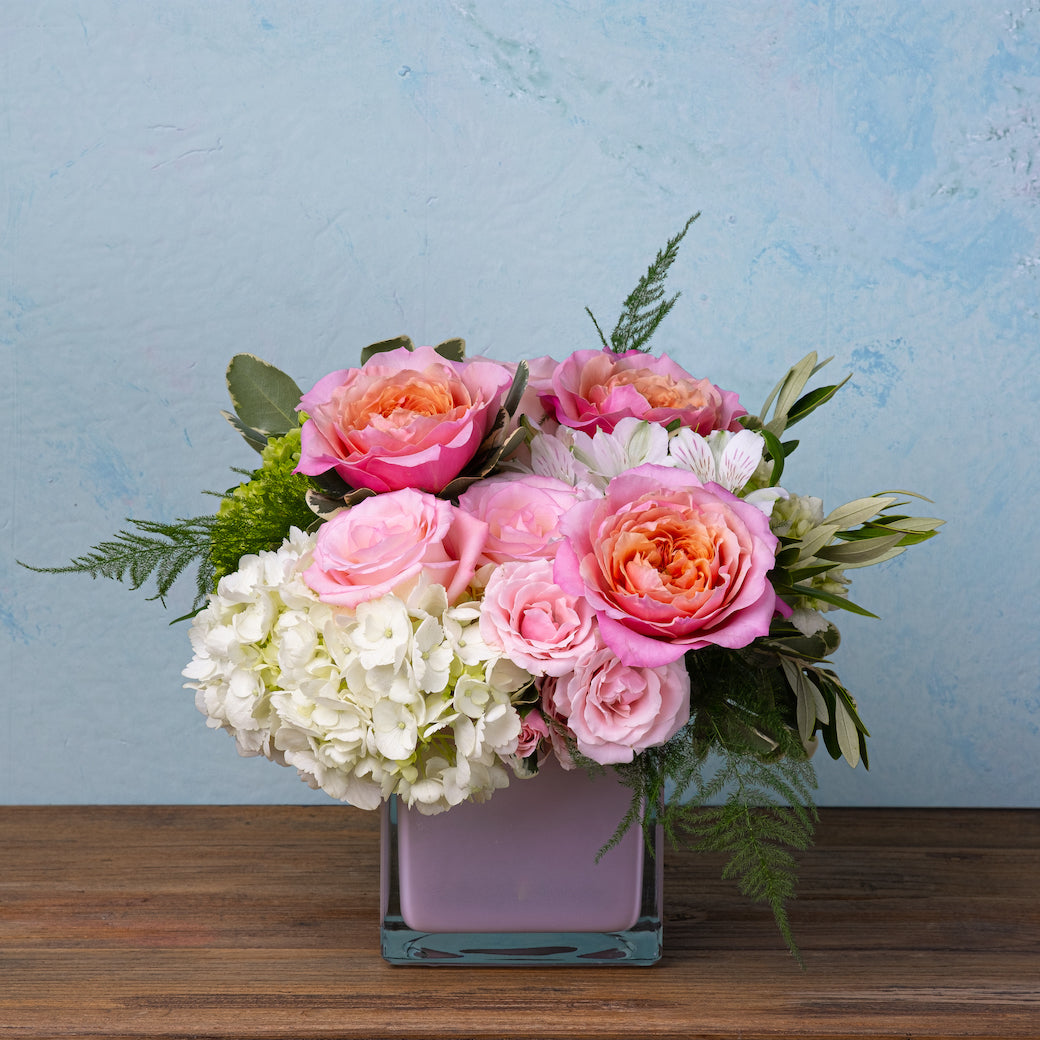 Square glass vase with pink roses, white hydrangeas, alstroemeria, and greenery in a compact floral arrangement.