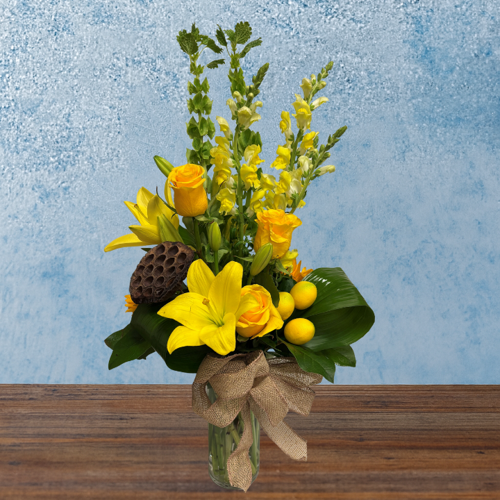 Tall arrangement of bright yellow flowers in a narrow cylindrical vase. Yellow lilies, roses and snap dragons accompany small lemons and a dried lotus pod. A burlap ribbon rests around the rim of the vase.