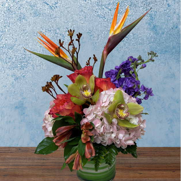 Foral arrangement in a cylindrical vase. Pink hydrangea, orchids and orange roses are densely clustered together. Birds of Paradise and Kangaroo Paw flowers complement the arrangement. 