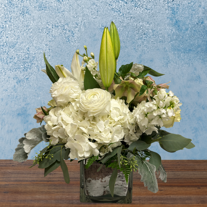 Floral arrangement in a cube glass vase. Flowers are white with lilies, roses, hydrangea and stock. Greenery supports the floral arrangement. 