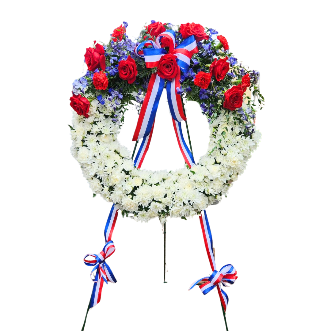 Patriotic floral wreath with red roses, white chrysanthemums, and blue accents, adorned with red, white, and blue ribbons, displayed on a stand.