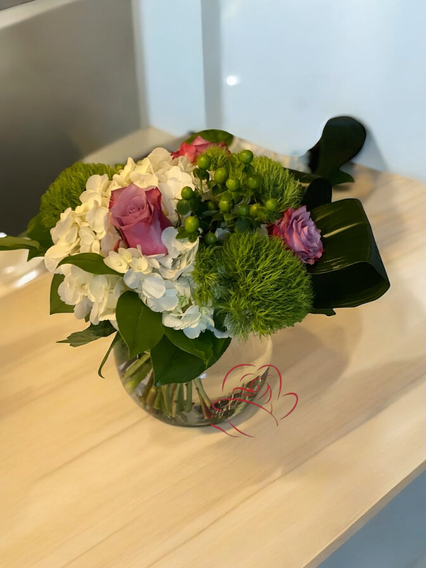 Chic floral arrangement with white hydrangeas, lavender roses, green dianthus, green hypericum berries, and lush foliage in a round glass vase on a wooden table.