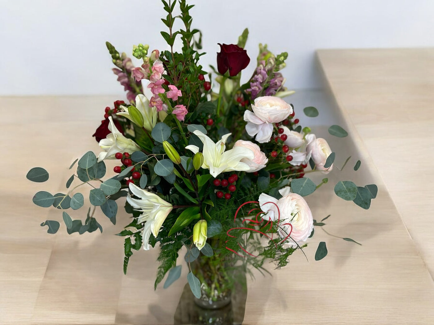 Elegant floral arrangement with white lilies, blush ranunculus, red roses, pink snapdragons, red berries, eucalyptus, and greenery in a clear glass vase, displayed on a wooden surface.