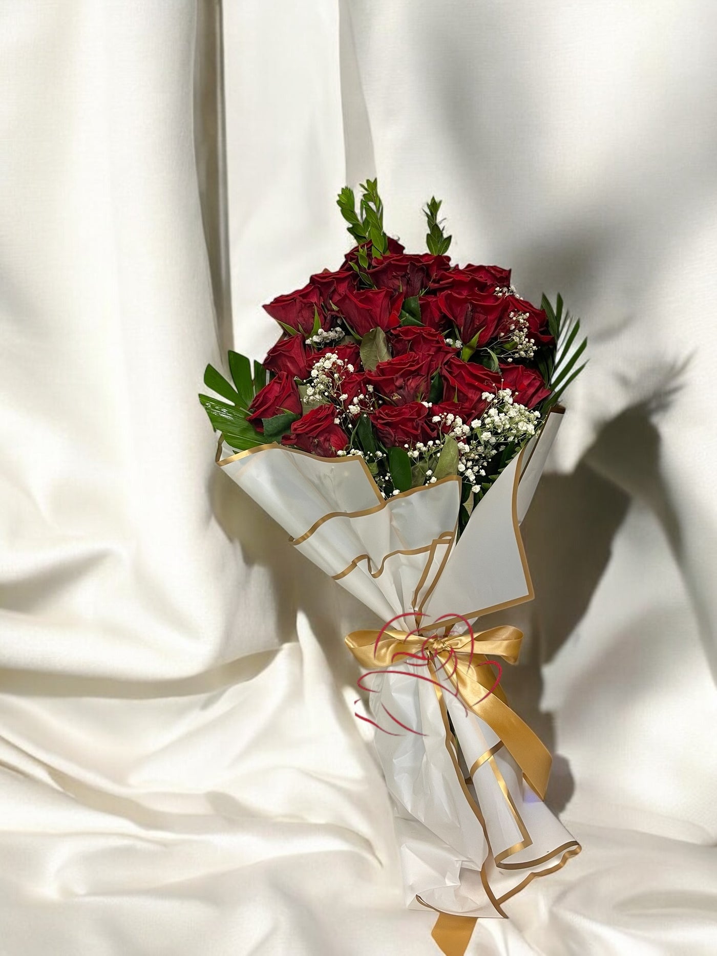 Elegant bouquet of red roses with baby's breath and greenery, wrapped in white paper with gold trim and tied with a gold satin ribbon, displayed against a white fabric background.