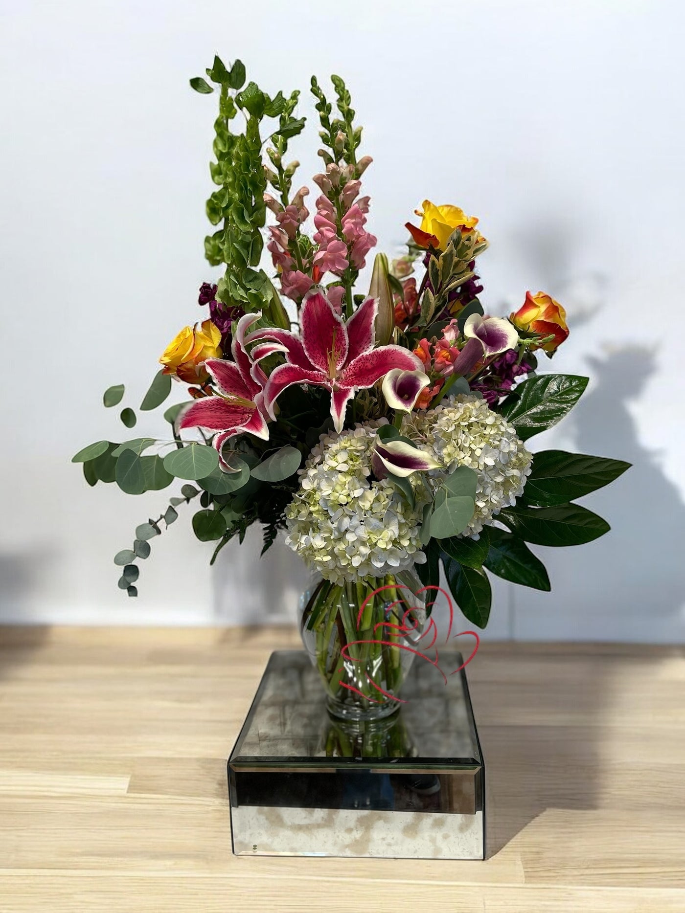 Mixed floral arrangement with pink Stargazer lilies, purple-edged calla lilies, yellow-orange roses, white hydrangeas, mauve snapdragons, and green foliage in a clear glass vase on a mirrored base