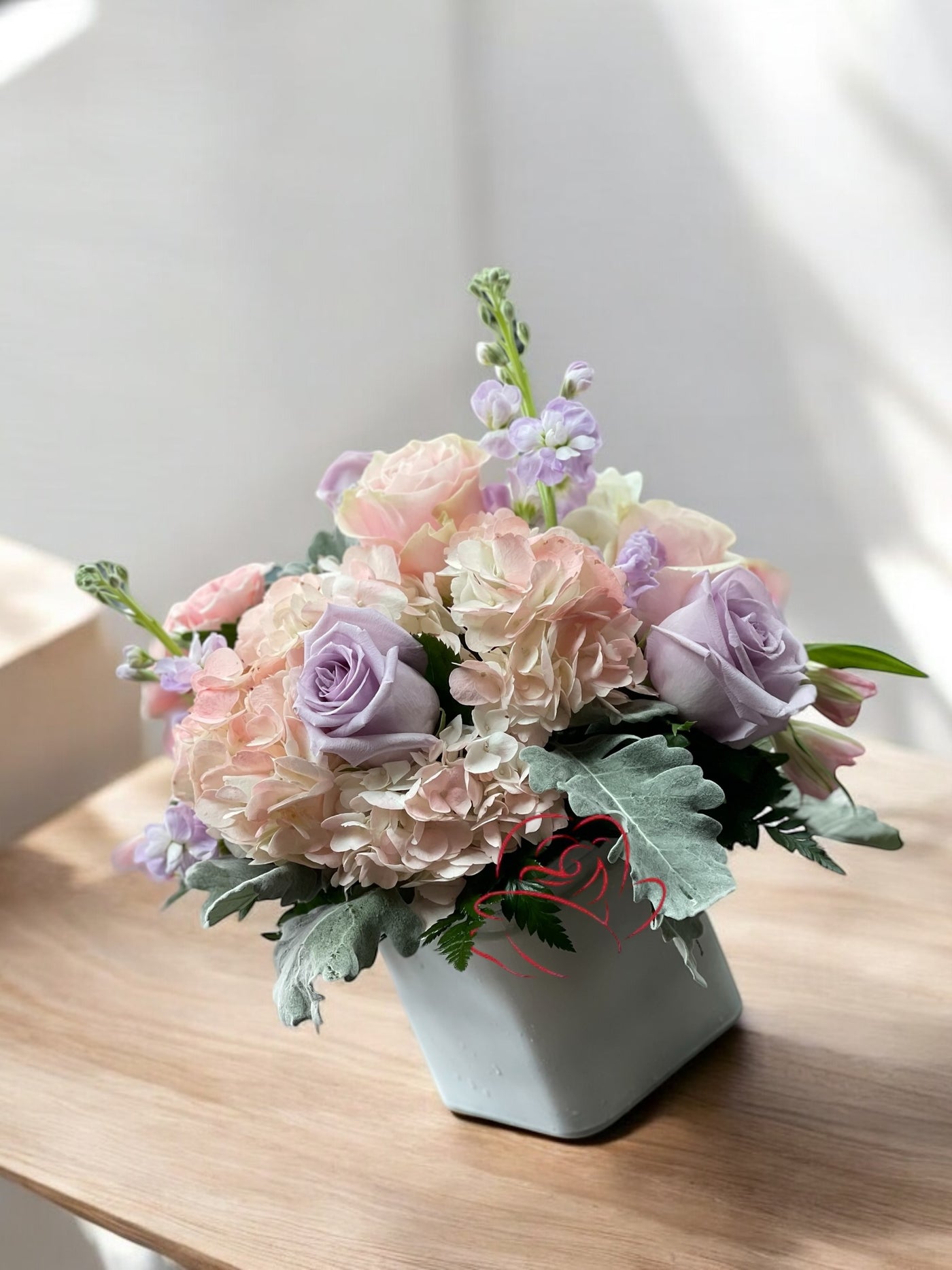 Pastel floral arrangement with lavender roses, blush hydrangeas, lilac stock flowers, and greenery in a white square vase, displayed on a wooden surface with sunlight streaming in.