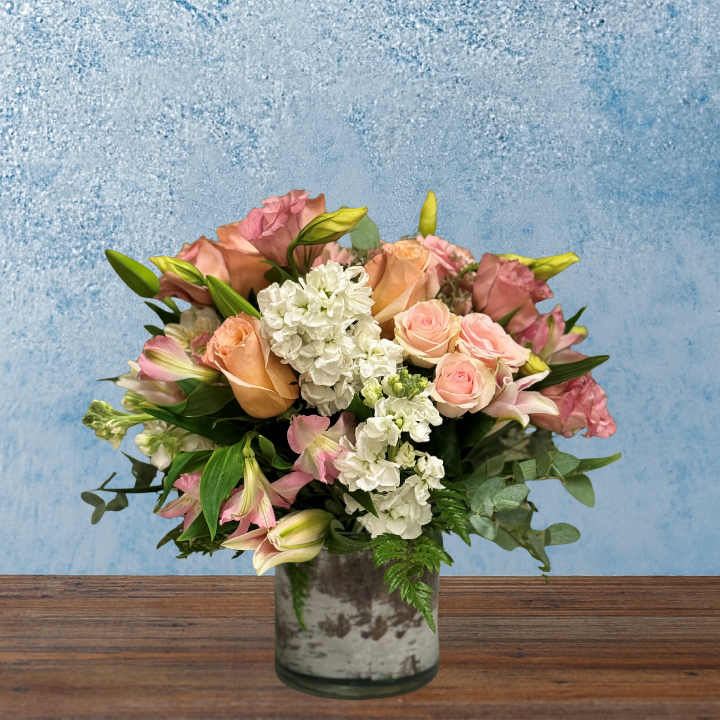 Densley packed floral arrangement in a cylindrical glass vase. Flowers are a mix of pastel pinks roses and alstroemeria with white stock. 