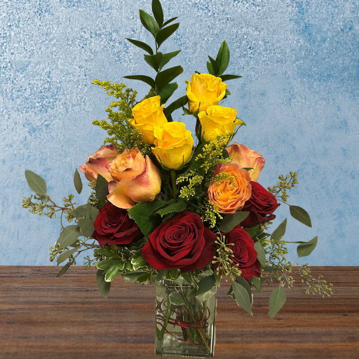 Tall arrangement with tiered roses in a rectangular vase. The bottom tier of roses are red, the middle are orange pink, and the top are yellow.