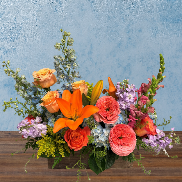 Vibrant and colorful arrangement in a rectangular wooden box, overflowing with flowers. Blue hydrangea, vibrant orange lilies and pink cabbage roses are accompanied by blue and purple stock stock, pink snapdragons and orange roses.