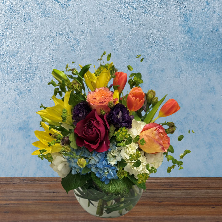 Colorful arrangement in a bubble bowl. A round arrangement consisting of yellow lilies, blue and white hydrangea, orange tulips, and a magenta rose.
