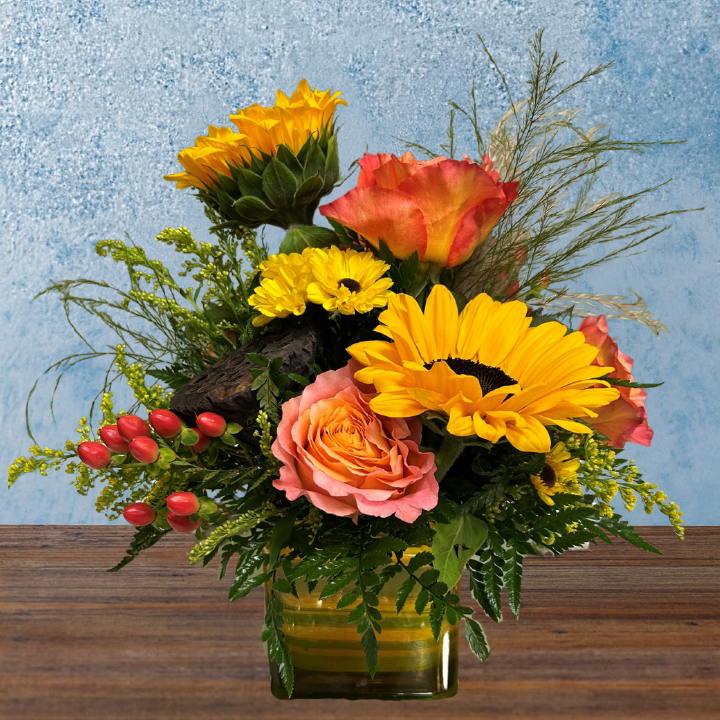 Flower arrangement in a cube vase. Visible flowers include, 2 yellow sunflowers, 3 pink-orange roses, a dried lotus pod and green foliage. 