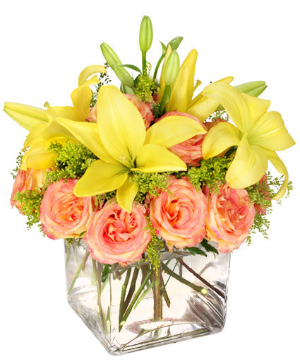 Floral arrangement with yellow lilies, peach roses, and greenery in a square glass vase.